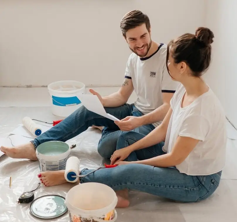 A couple sits on the floor surrounded by paint and brushes, planning their home improvement project with enthusiasm